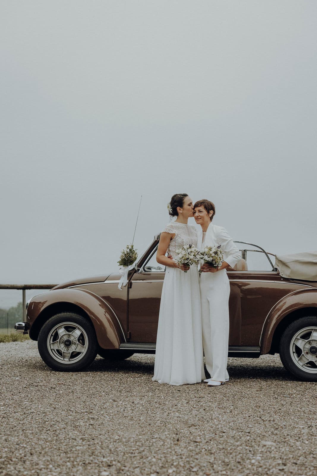 Zwei Bräute mit Brautsträußen vor einem braunen VW-Käfer-Oldtimer