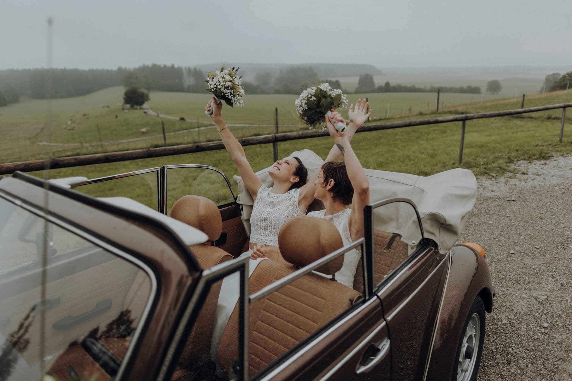 Braut jubelt mit erhobenem Brautstrauß im offenen Oldtimer vor nebliger Landschaft
