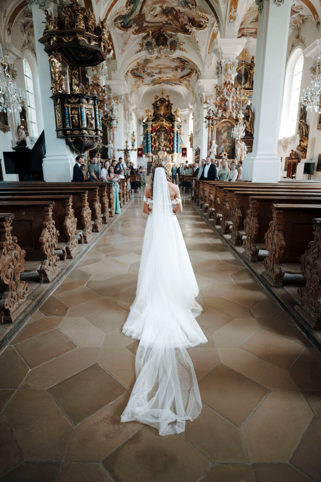 Braut schreitet mit langem Schleier durch das Mittelschiff einer barocken Kirche