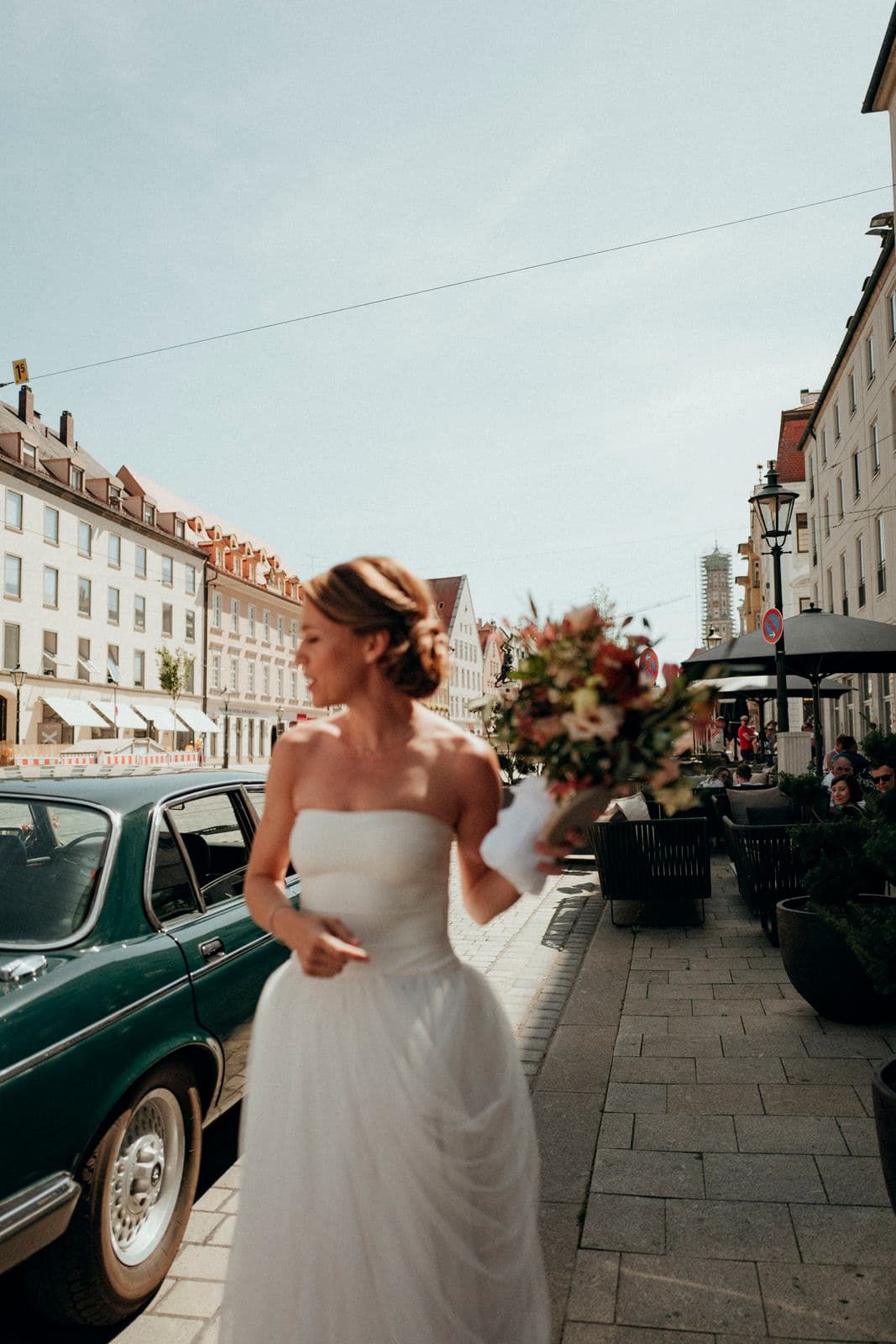Braut mit Brautstrauß auf einer Stadtstraße neben einem grünen Oldtimer