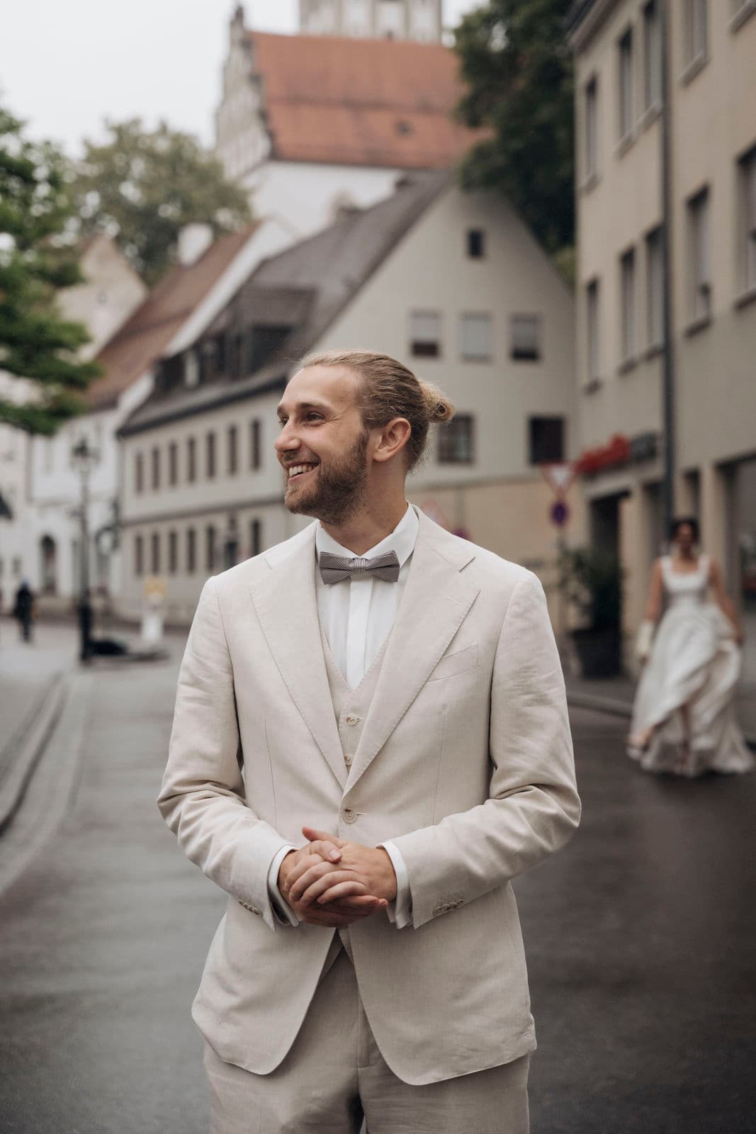 Bräutigam im hellen Leinenanzug wartet lächelnd in der Altstadtgasse, im Hintergrund nähert sich die Braut zum First Look
