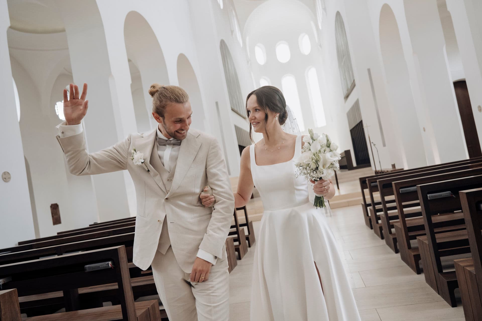 Brautpaar zieht Arm in Arm durch den Mittelgang der lichtdurchfluteten weißen Kirche, der Bräutigam winkt den Gästen zu