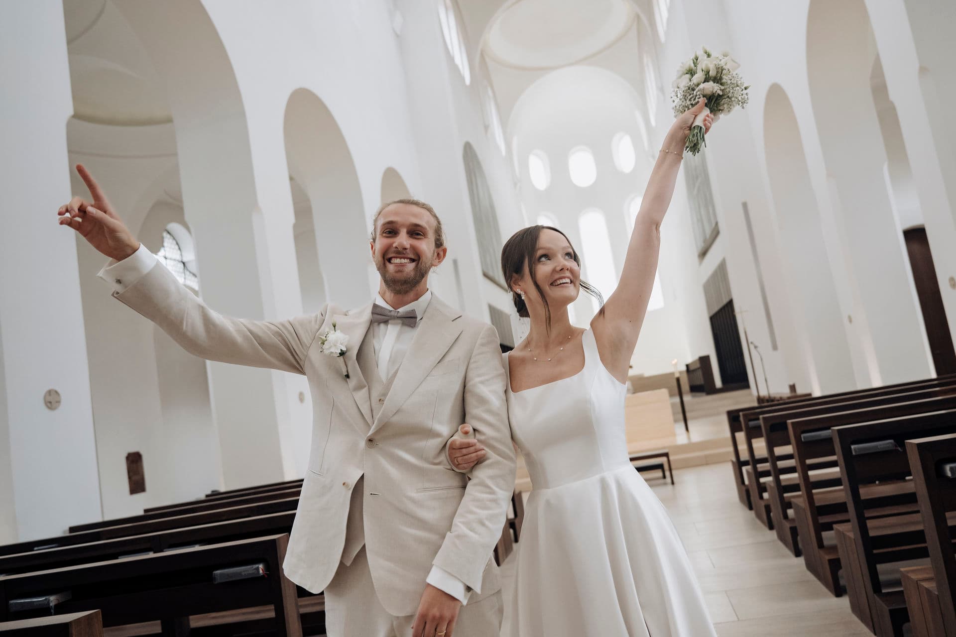 Brautpaar jubelt beim Auszug in der weißen Kirche, die Braut reckt den Brautstrauß in die Höhe