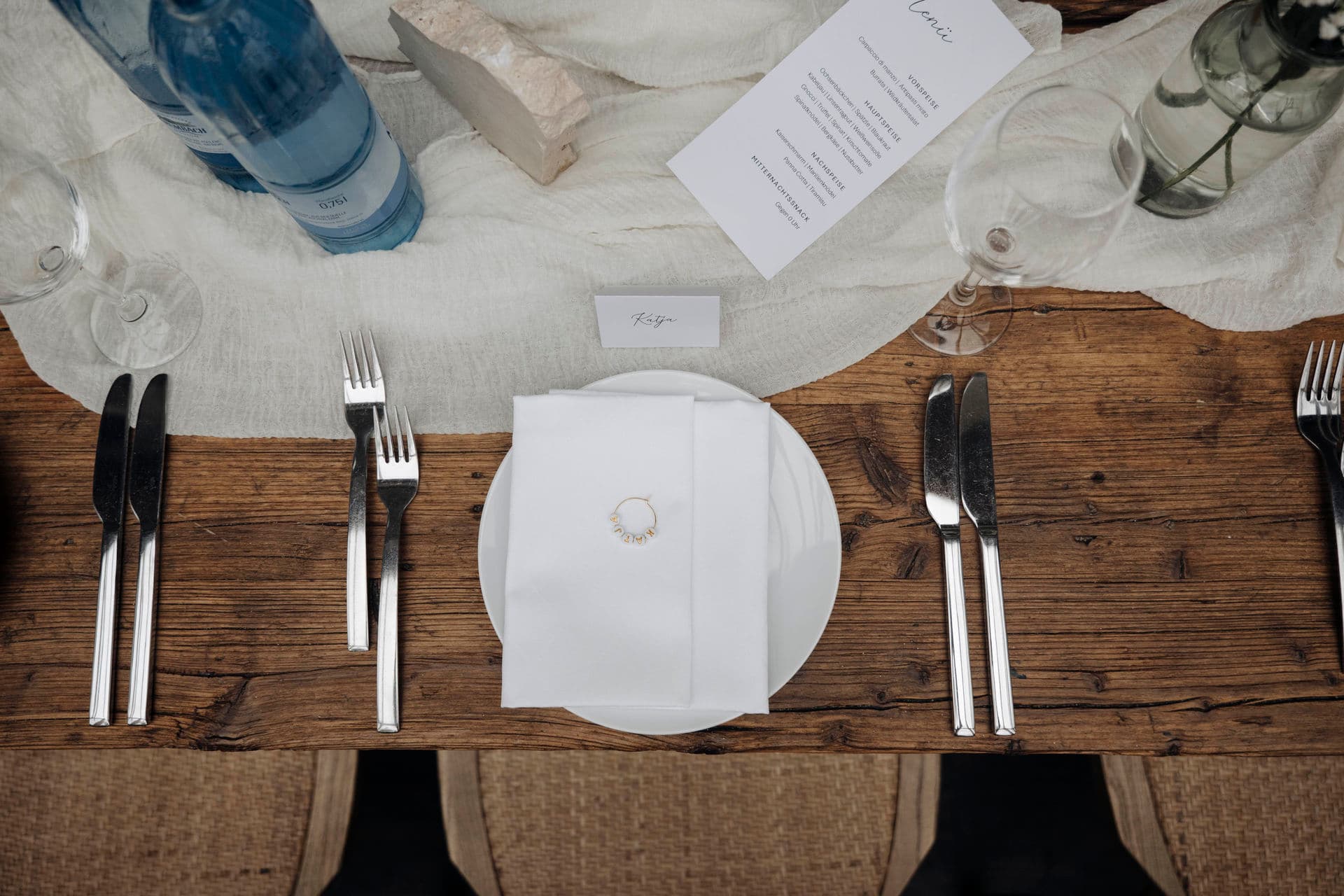 Gedeck von oben: weiße Serviette mit Glasring, Platzkarte und Menükarte auf dem rustikalen Holztisch