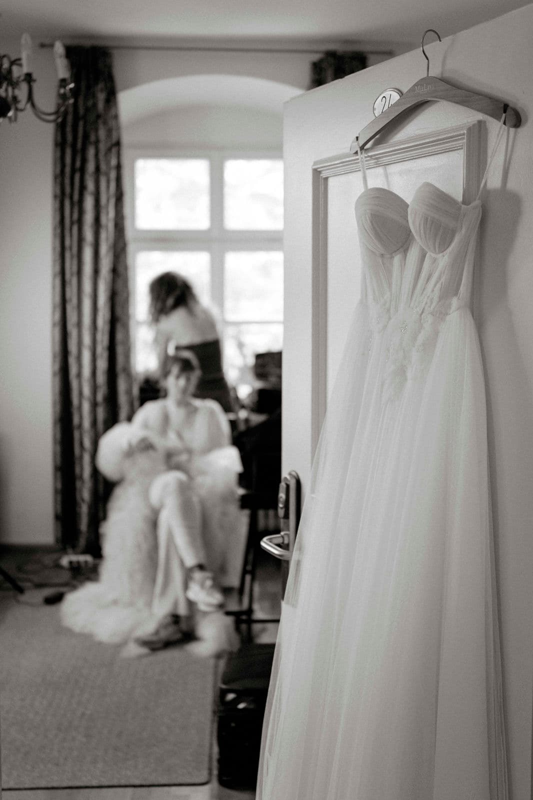 Brautkleid aus Tüll hängt am Türrahmen, im Hintergrund die Braut beim Getting Ready – Schwarz-Weiß