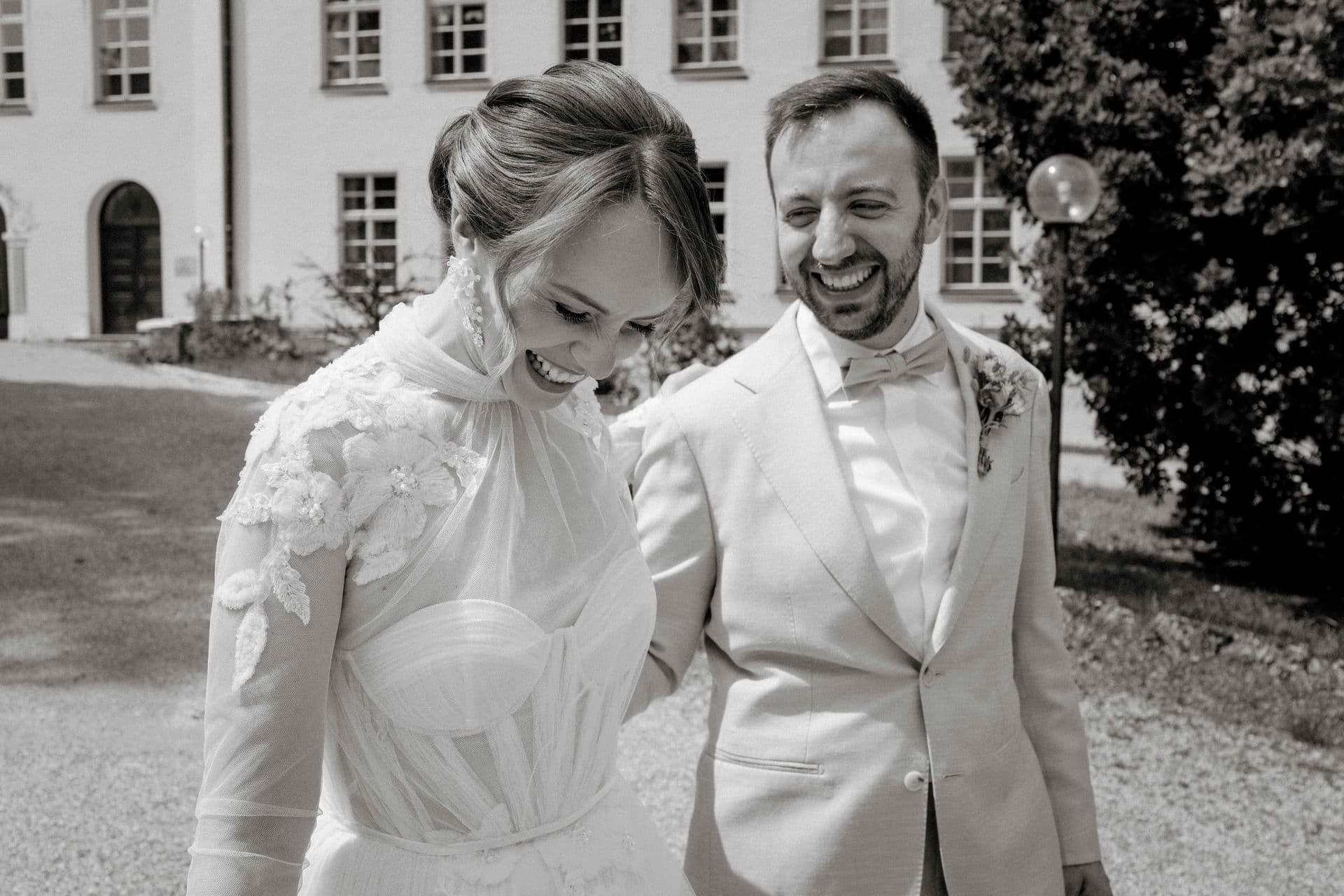 Brautpaar lacht beim Spaziergang vor dem Schloss, der Bräutigam im hellen Anzug mit Fliege – Schwarz-Weiß-Reportage