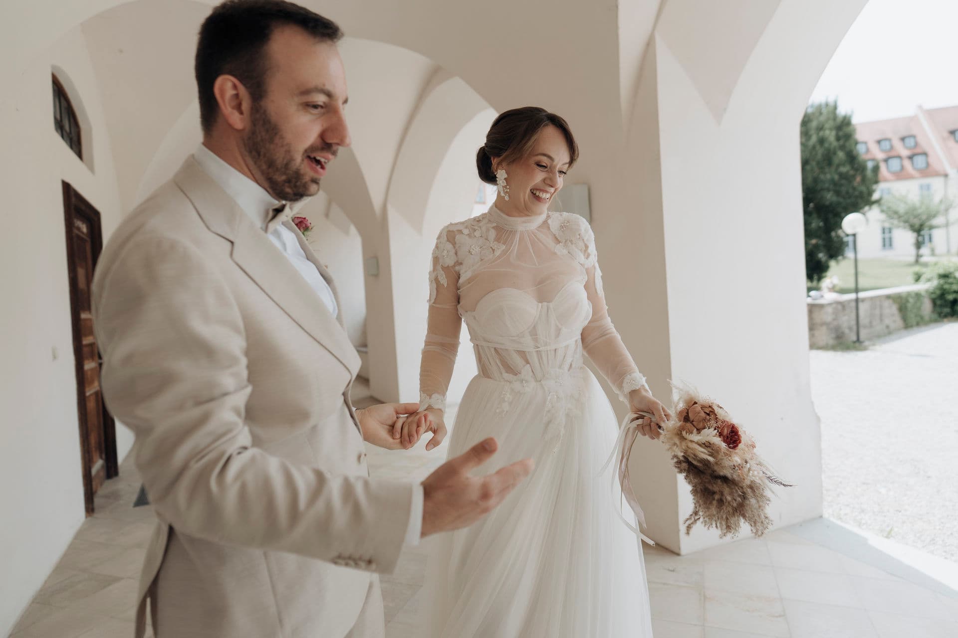 Brautpaar Hand in Hand im Gewölbegang, die Braut mit Trockenblumenstrauß im Spitzenkleid