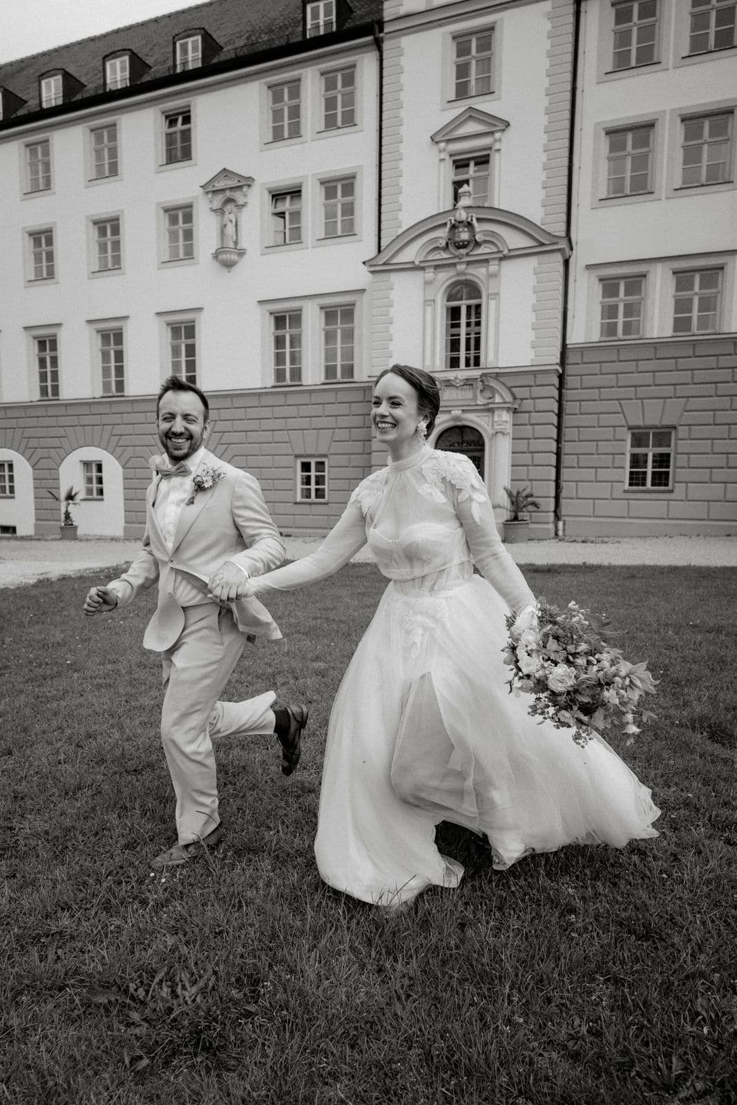 Brautpaar läuft ausgelassen Hand in Hand über den Rasen, das Tüllkleid der Braut weht im Schritt – Schwarz-Weiß