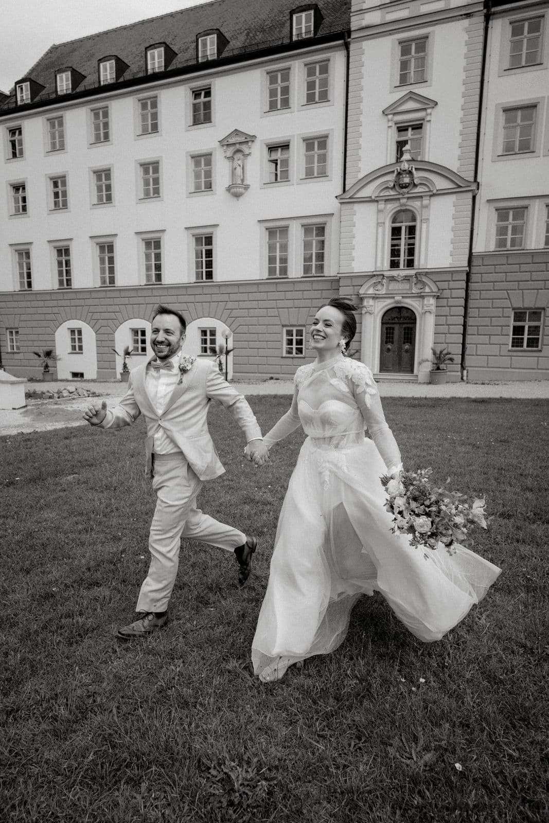 Lachendes Brautpaar rennt Hand in Hand vor dem barocken Schlossportal über die Wiese – Schwarz-Weiß-Reportage