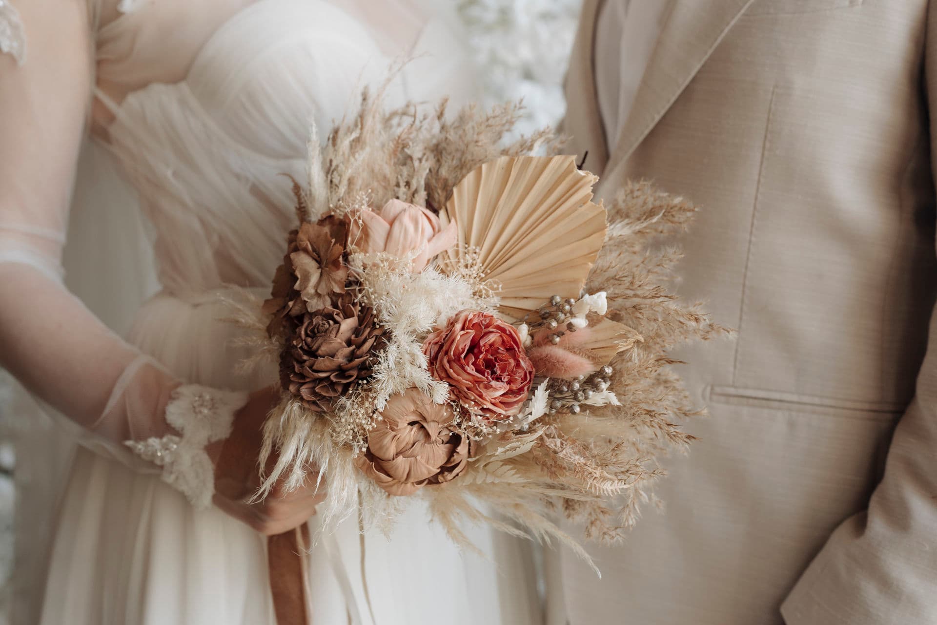 Nahaufnahme des Boho-Brautstraußes aus Trockenblumen vor Brautkleid und beigem Anzug