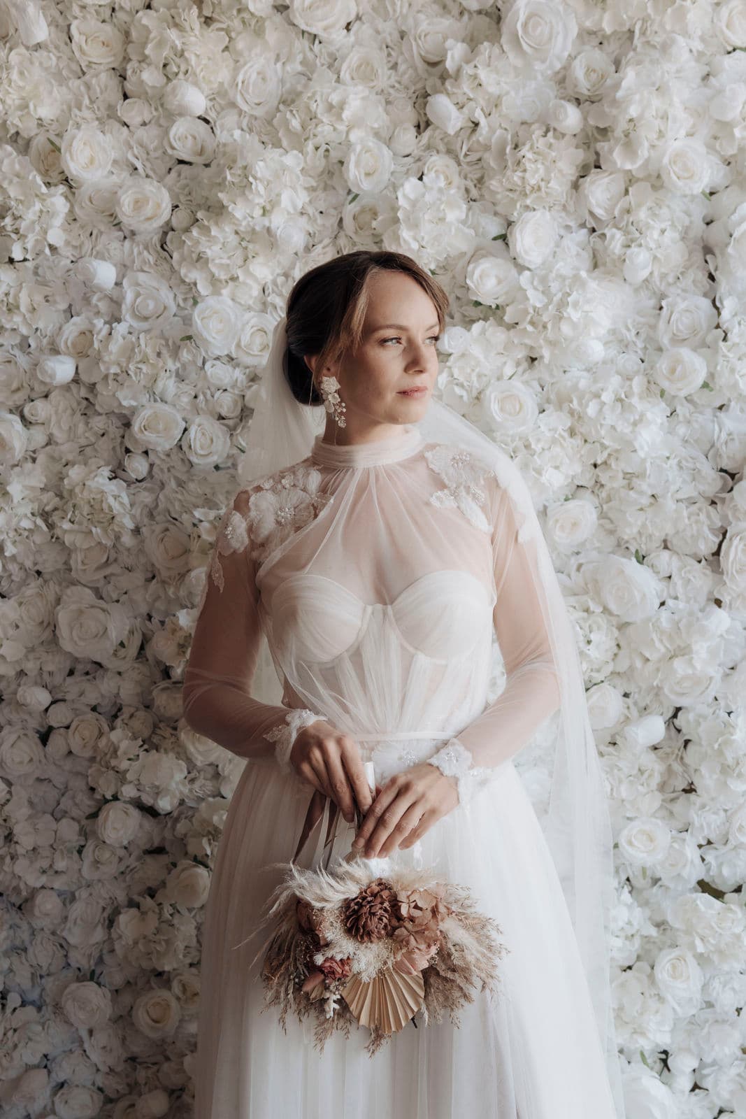 Braut mit Schleier und transparentem Tüllkleid blickt vor der weißen Rosenwand zur Seite