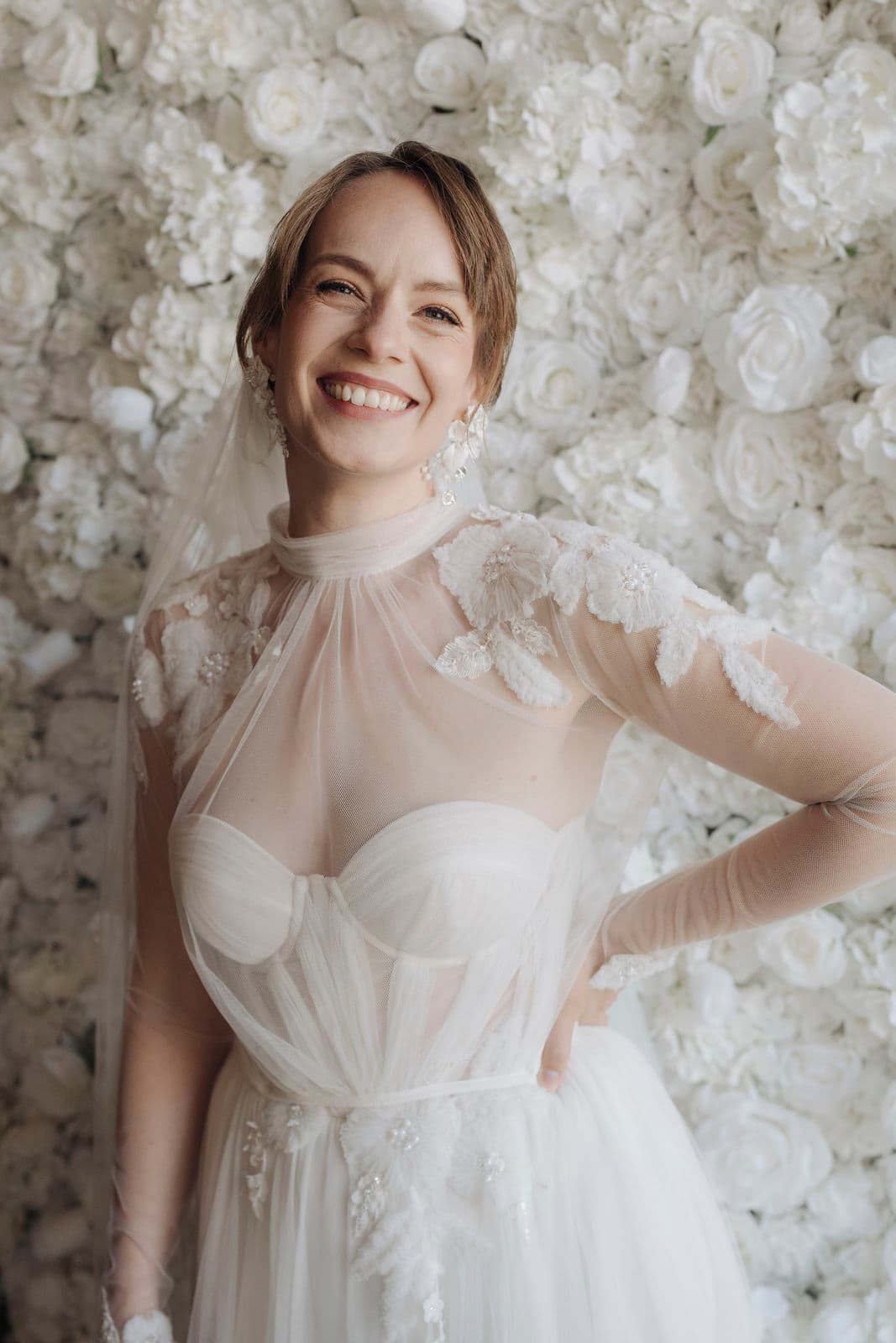 Strahlend lachende Braut im Tüllkleid mit Blütenapplikationen vor der weißen Blumenwand