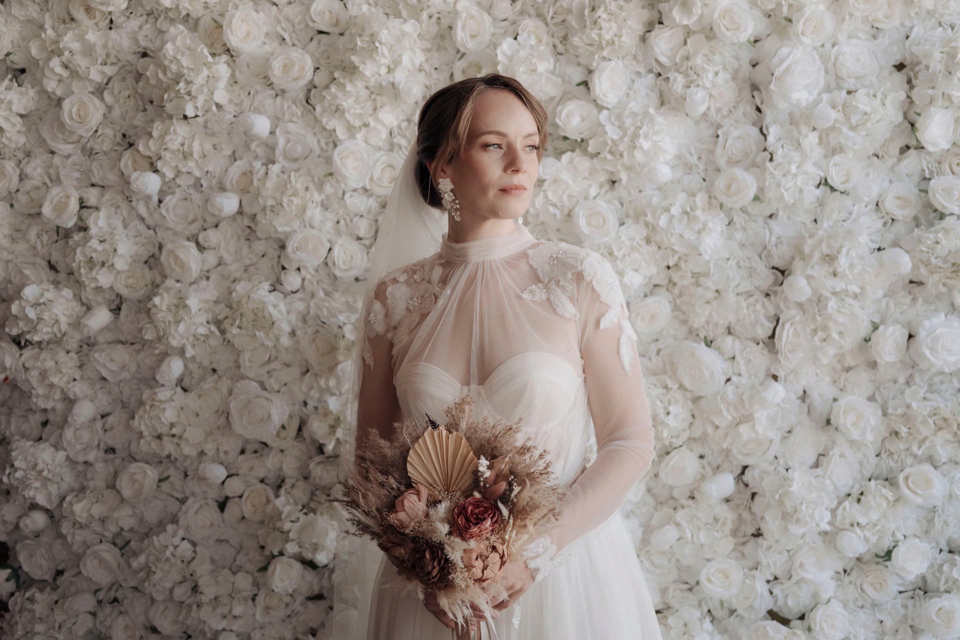 Braut steht mit Brautstrauß vor der raumhohen weißen Blumenwand und blickt ins Licht