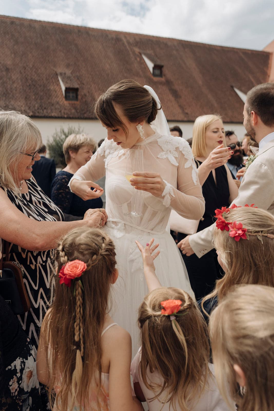 Braut inmitten gratulierender Gäste und Blumenmädchen mit Blüten im Haar beim Sektempfang im Innenhof